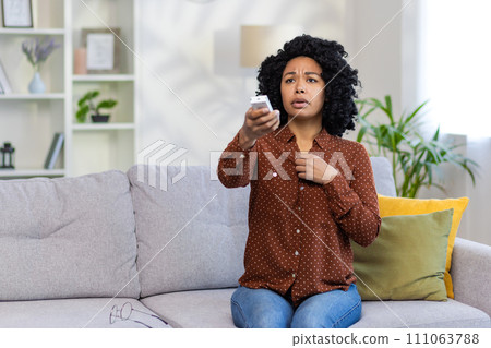 Worried young African American woman sitting on sofa at home and turning on air conditioner by remote control. He feels bad. 111063788