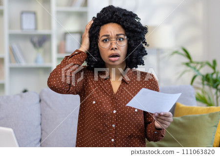 Worried young woman with glasses holding a document, expressing stress and confusion at home, concept of financial or personal crisis. Worried young woman with glasses holding a document, expressing stress and confusion at home, concept of financial or personal crisis. 111063820