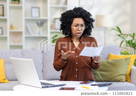 Shocked african american woman sitting on sofa at home, working with laptop and documents and looking upset at received bills and letter. Shocked african american woman sitting on sofa at home, working with laptop and documents and looking upset at received bills and letter. 111063821
