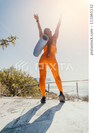 Fitness woman sea. Happy middle aged woman in orange sportswear exercises morning outdoors on yoga mat with laptop in park over ocean beach. Female fitness pilates yoga routine. Healthy lifestyle. 111065132