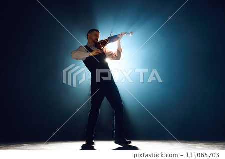 Classical musician performing on violin, playing melody on stage with spotlight from behind him surrounded smoke. Classical musician performing on violin, playing melody on stage with spotlight from behind him surrounded smoke. 111065703