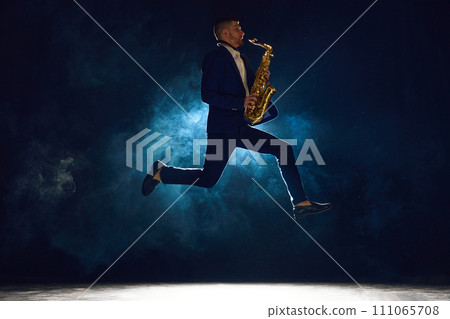 Dynamic shot of artistic man, solo performer jumping while playing jazz melody on saxophone against dark background with smoke. Dynamic shot of artistic man, solo performer jumping while playing jazz melody on saxophone against dark background with smoke. 111065708