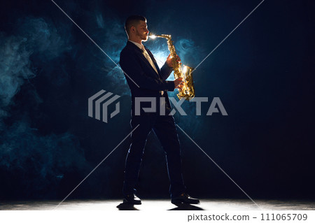 Solo saxophonist in concert outfit performing with intense expression on stage with backlight against dark background with dramatic smoke. Solo saxophonist in concert outfit performing with intense expression on stage with backlight against dark background with dramatic smoke. 111065709