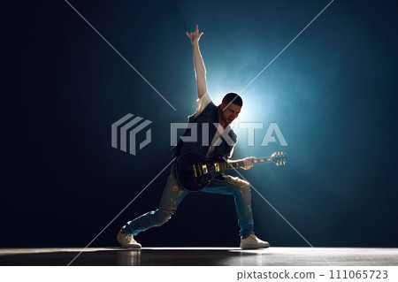 Guitar player engaging with music and shows Rock-n-Roll gesture on stage with backlights against black background with smoke. Guitar player engaging with music and shows Rock-n-Roll gesture on stage with backlights against black background with smoke. 111065723
