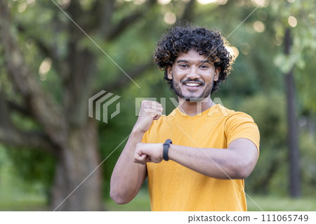 Smiling young Indian athlete enjoying a workout in the park, pausing to check progress on his smartwatch. Smiling young Indian athlete enjoying a workout in the park, pausing to check progress on his smartwatch. 111065749