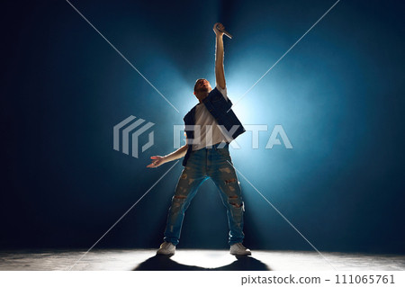 Street style singer gesturing with hand on stage against dark background. Spotlight and smoke creating silhouette effect. 111065761