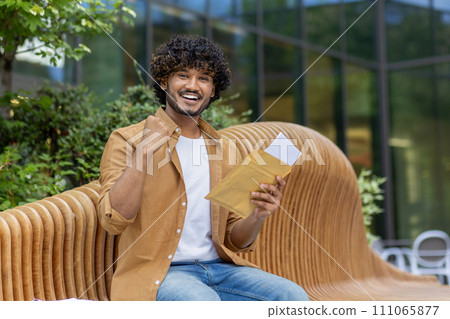 Portrait of a young Muslim man sitting on a bench in the street, holding an open envelope with a letter, smiling and happy to the camera, showing a victory gesture with his hand. Portrait of a young Muslim man sitting on a bench in the street, holding an open envelope with a letter, smiling and happy to the camera, showing a victory gesture with his hand. 111065877