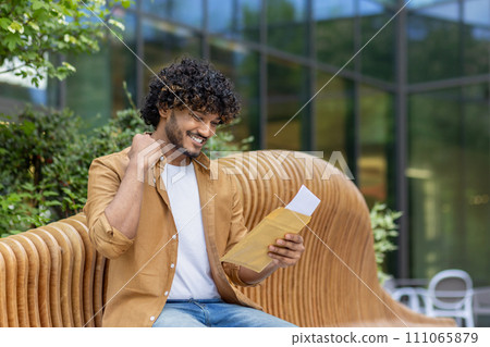 Indian young man received good news, sitting on a bench outside, reading a letter and happy with a victory gesture with his hand. Indian young man received good news, sitting on a bench outside, reading a letter and happy with a victory gesture with his hand. 111065879