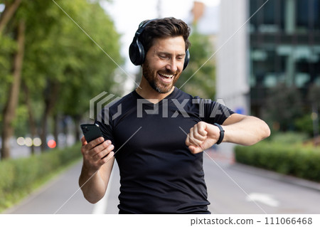 A happy young man is standing on a city street wearing headphones, holding a phone in his hand and looking contentedly at a smart watch. Close-up photo. 111066468