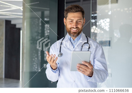 A young smiling male doctor is standing in the hospital lobby in a white coat and talking via video communication with the patient via a tablet. He explains, consults. A young smiling male doctor is standing in the hospital lobby in a white coat and talking via video communication with the patient via a tablet. He explains, consults. 111066636