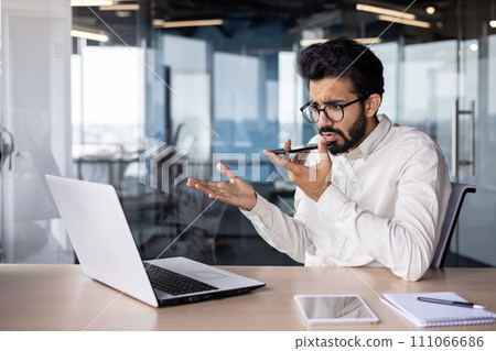 Serious young Indian male businessman sitting in the office at the desk and talking emotionally on the phone through the loudspeaker, using voice search and recording the conversation. 111066686