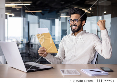 Muslim young man is happy with the received letter and documents with results, sitting in the office at the table, reading the message, showing a victory gesture with his hand. Muslim young man is happy with the received letter and documents with results, sitting in the office at the table, reading the message, showing a victory gesture with his hand. 111066694