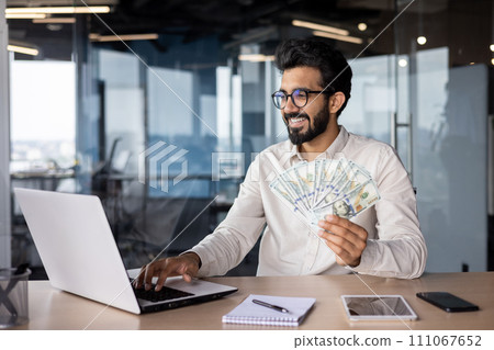 Indian smiling young man businessman working in the office on a laptop, sitting at the table and holding cash bills in his hand. 111067652