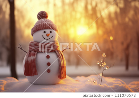 Funny snowman in a red scarf and hat on the snow in the park in the rays of the setting sun. 111070578