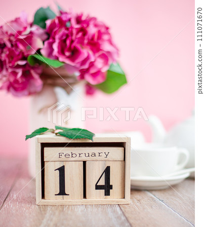 Wooden calendar with Valentine's Day for February 14 on the background of flowers and a cup of hot drink for breakfast 111071670