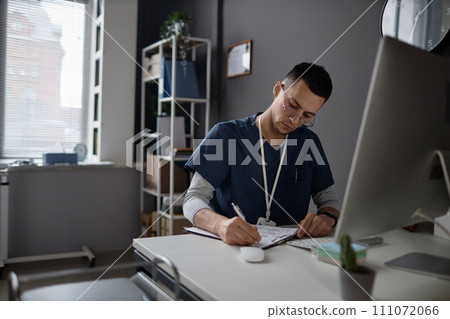 Medium shot of focused Islamic man doctor wearing eyeglasses and scrubs doing medical paperwork sitting at table in hospital Medium shot of focused Islamic man doctor wearing eyeglasses and scrubs doing medical paperwork sitting at table in hospital 111072066