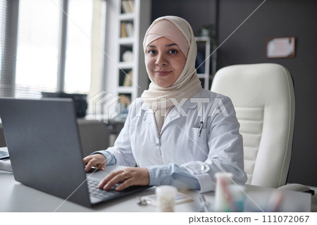 Portrait of friendly Islamic female health practitioner in hijab looking at camera while working on laptop sitting at desk in modern clinic 111072067