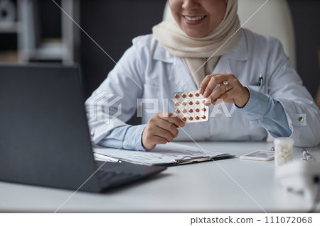 Cropped shot of smiling female doctor wearing hijab showing blister pack of pills to patient at video call using laptop in hospital 111072068
