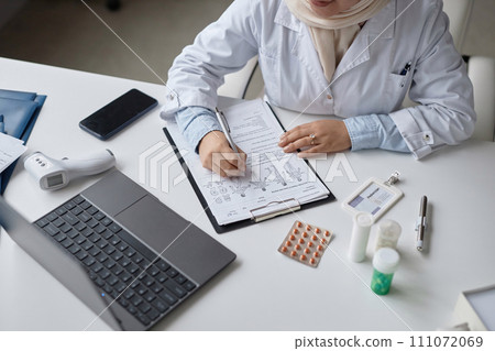 Top down view of unrecognizable female physician in headscarf filling in medical questionnaire at online appointment with patient sitting at workplace in clinic 111072069