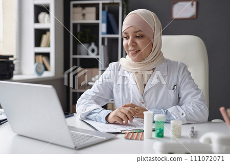 Medium shot of cheerful Muslim woman physician in beige headscarf consulting patient remotely using laptop sitting at clinic desk 111072071