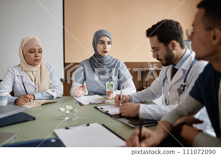 Experienced Muslim woman doctor in gray hijab giving speech during staff training while sitting at desk in meeting room Experienced Muslim woman doctor in gray hijab giving speech during staff training while sitting at desk in meeting room 111072090