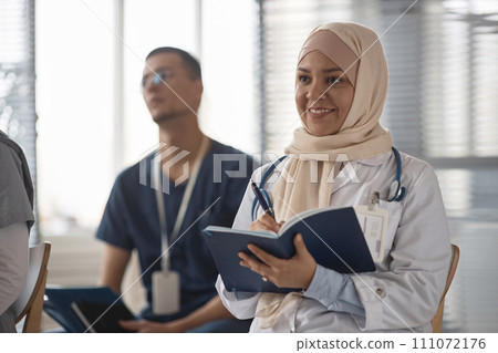 Medium shot of cheerful Muslim woman doctor in beige headscarf holding notebook while listening to presentation at conference Medium shot of cheerful Muslim woman doctor in beige headscarf holding notebook while listening to presentation at conference 111072176