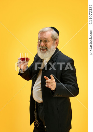 Portrait of positive, smiling senior man holds glass of whiskey and pointing to camera against yellow background. Happy Purim. 111072322