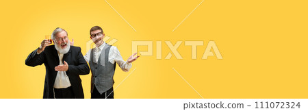 Banner. Two positive young and senior men posing in formal suits against yellow background with negative space to insert text. 111072324