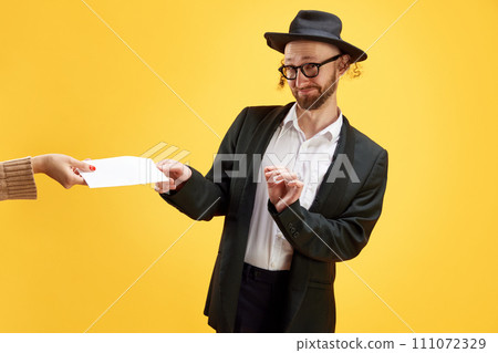 Skeptical Jewish man in hat and formal suit receiving envelope from hand against bright yellow background. Culture and business. Skeptical Jewish man in hat and formal suit receiving envelope from hand against bright yellow background. Culture and business. 111072329