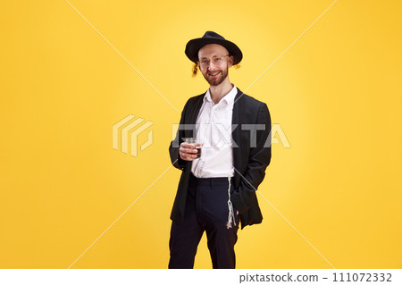 Confident man in formal Jewish attire, holding glass of alcohol drink and looking at camera against warm yellow studio background. Confident man in formal Jewish attire, holding glass of alcohol drink and looking at camera against warm yellow studio background. 111072332