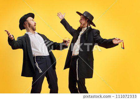 Two Jewish men in traditional attire, one guy with wine glass and other with colored wooden grogger against yellow background. Two Jewish men in traditional attire, one guy with wine glass and other with colored wooden grogger against yellow background. 111072333