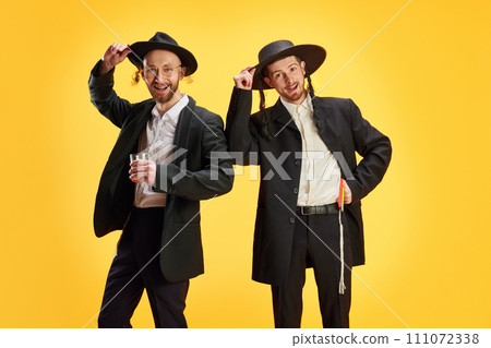 Jewish men in traditional suit and hats posing holds glass of alcohol drinks against vivid yellow background. Jewish men in traditional suit and hats posing holds glass of alcohol drinks against vivid yellow background. 111072338