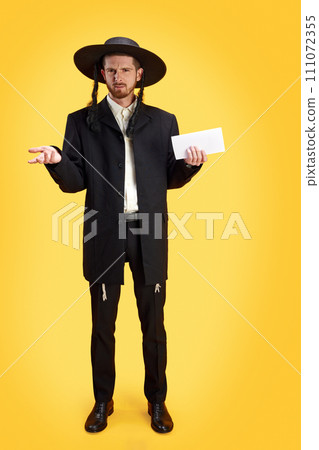 Skeptical Jewish man in traditional garb holding an envelope, evoking curiosity and intrigue against sunny yellow background. 111072355