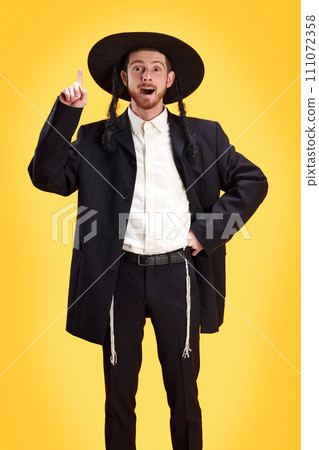 Portrait of young Jewish man holds one hand raised up while he create a new idea against sunny yellow background. Portrait of young Jewish man holds one hand raised up while he create a new idea against sunny yellow background. 111072358