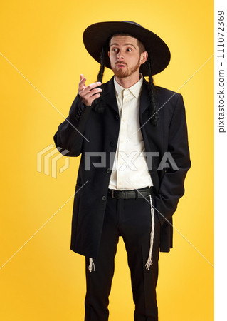 Pensive man in traditional Jewish attire with wide-brimmed hat and side curls posing against sunny yellow background. 111072369