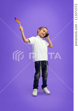 Little boy dressed modern casual outfit playing with grogger toy and talking on phone against purple studio background. Little boy dressed modern casual outfit playing with grogger toy and talking on phone against purple studio background. 111072372