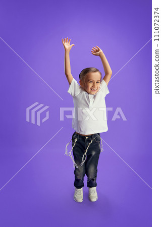 Ecstatic child wearing white shirt and jeans with raised hands expressing joy and happiness against purple studio background. Ecstatic child wearing white shirt and jeans with raised hands expressing joy and happiness against purple studio background. 111072374