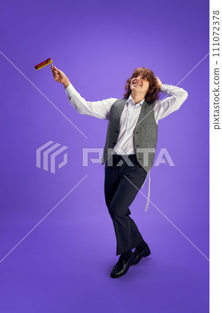 Portrait of happy young Jewish man in formal suit and kippah dancing with grogger against purple studio background. 111072378