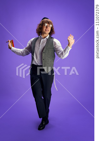 Young Jewish man with grogger dancing national dance holds grogger against purple studio background. Purim holiday. Ad 111072379