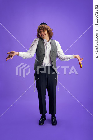 Cheerful man in tweed waistcoat and kippah with arms outstretched, holding grogger against purple studio background. Cheerful man in tweed waistcoat and kippah with arms outstretched, holding grogger against purple studio background. 111072382