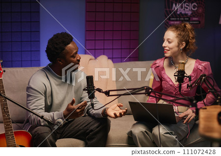 Young Black male guitarist talking to stylish smiling Caucasian woman with laptop during podcast sitting on sofa on studio 111072428