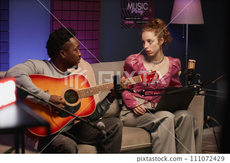 African American male musician playing acoustic guitar sitting on couch with young Caucasian female podcaster holding laptop in studio with neon light 111072429