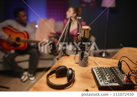 Professional microphones, headphones and sound mixer on desktop in podcast room with unrecognizable music bloggers in blurred background 111072430
