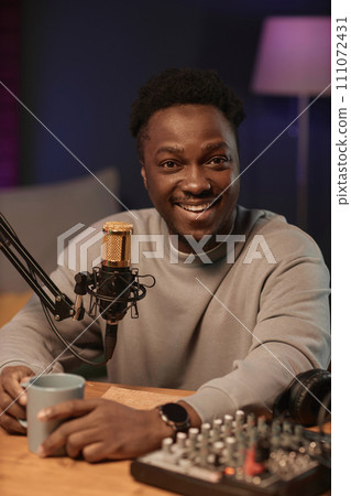 Portrait of happy Black man looking at camera holding cup sitting at table with audio equipment in podcast studio Portrait of happy Black man looking at camera holding cup sitting at table with audio equipment in podcast studio 111072431