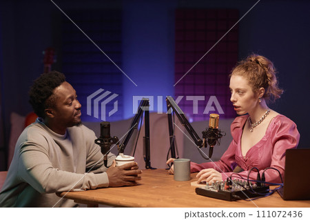 Medium shot of Black man listening to young Caucasian female podcast host at table with studio equipment and coffee cups in room with neon light Medium shot of Black man listening to young Caucasian female podcast host at table with studio equipment and coffee cups in room with neon light 111072436