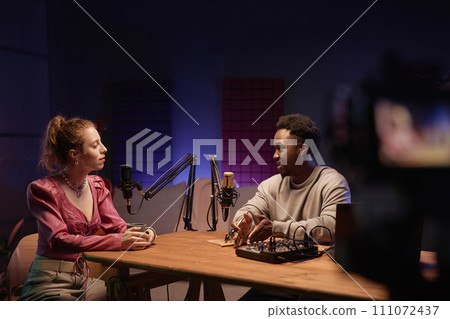 Two friendly diverse bloggers at desk in comfy neon lit studio discussing issues while recording podcast on camera Two friendly diverse bloggers at desk in comfy neon lit studio discussing issues while recording podcast on camera 111072437