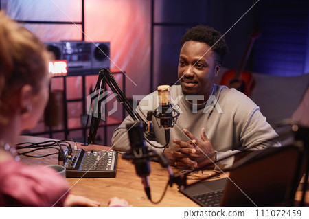 Young African American man carefully listening to unrecognizable woman sitting at desk with studio equipment int podcast room with neon light Young African American man carefully listening to unrecognizable woman sitting at desk with studio equipment int podcast room with neon light 111072459