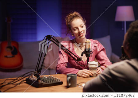 Smiling Caucasian girl with cup listening to unrecognizable man during podcast while sitting at table with microphone and sound mixer in neon light studio 111072462