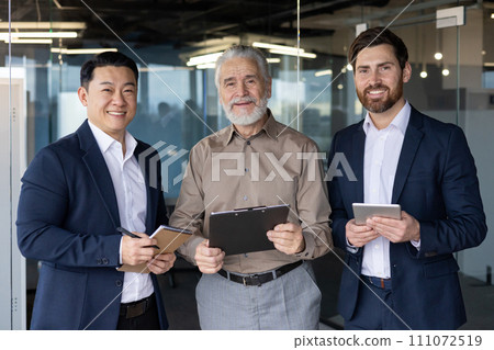 Three diverse professional businessmen smiling and standing together in an office setting, showcasing teamwork and collaboration. 111072519