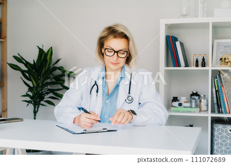 Confident mature middle aged woman doctor medical nurse, female physician practitioner in white coat with stethoscope sitting at working place at hospital office and making notes during appointment. Confident mature middle aged woman doctor medical nurse, female physician practitioner in white coat with stethoscope sitting at working place at hospital office and making notes during appointment. 111072689